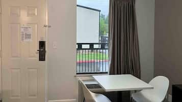 A hotel room with a white door, a window with a view of a balcony, a small table, and a white chair.