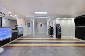En hotelllobby med en receptionsdisk, en digital skärm, en bankomat och en trappa med en färgglad rand.