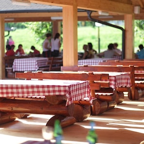 Un espace extérieur abrité, aménagé pour les repas, avec des tables et des bancs en bois, dont certaines sont recouvertes de nappes à carreaux.