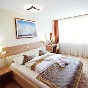 Une chambre d’hôtel avec un lit, un bureau, une chaise, un tableau au mur et une fenêtre avec des rideaux.