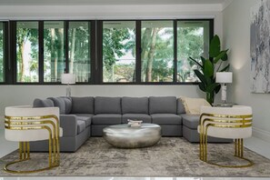 A modern living room with a grey sectional sofa, a round coffee table, and two white chairs with gold accents. Large windows offer a view of greenery outside. - 8,000 sq ft Estate with Heated Pool & Game Room (Plantation)