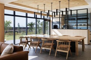 A modern dining area with a wooden table and chairs, large windows, and a kitchen area with a herringbone-patterned backsplash.