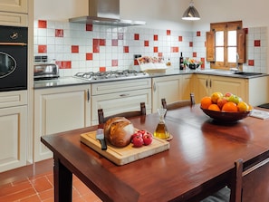 Una cocina con gabinetes blancos, salpicadero de azulejos, estufa a gas y una mesa de comedor de madera con un tazón de fruta.
