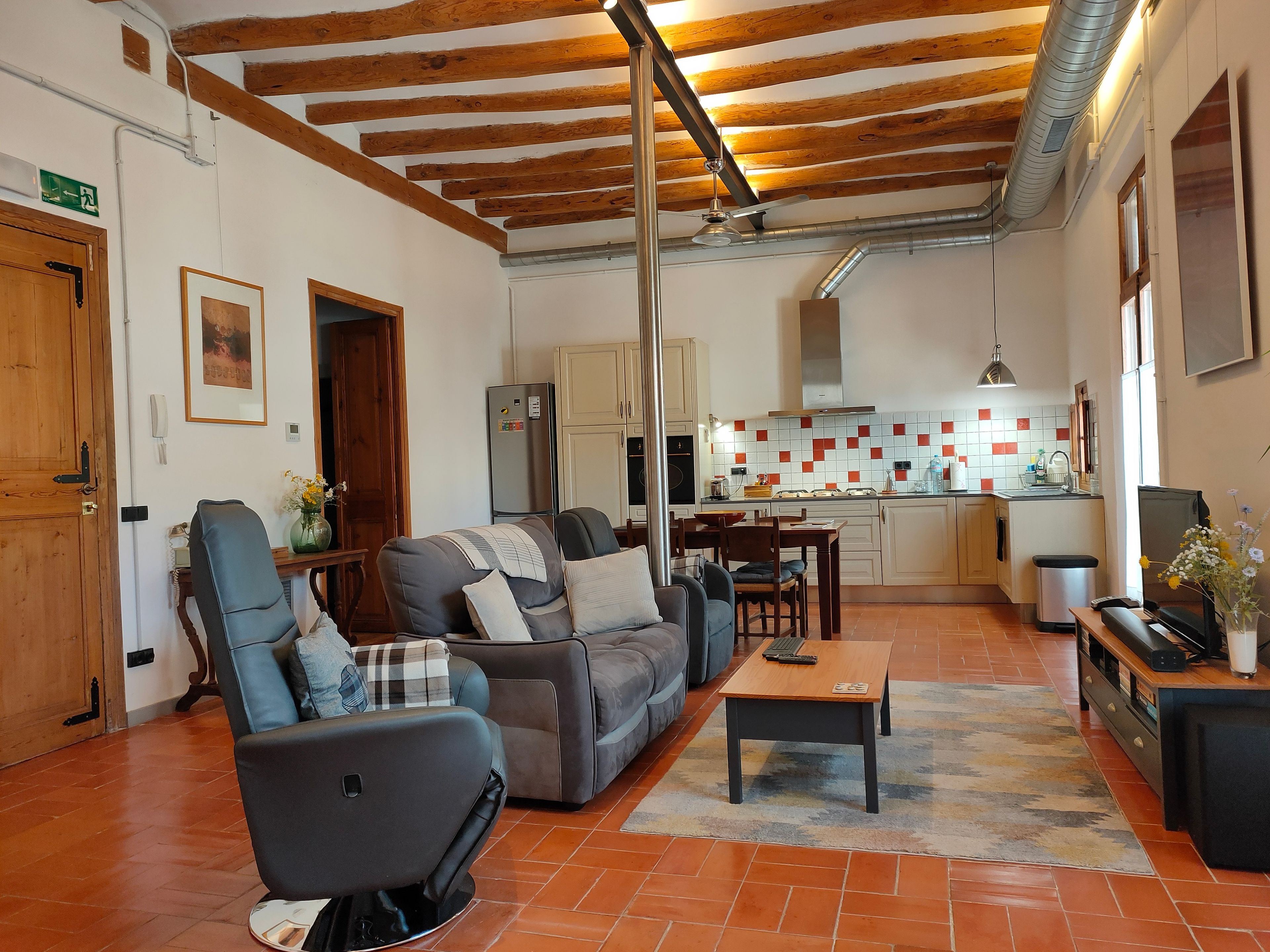 Ampla sala de estar com sofá, poltrona e mesa de centro. Uma cozinha com painel de azulejos e uma área de jantar com mesa e cadeiras são visíveis ao fundo.
