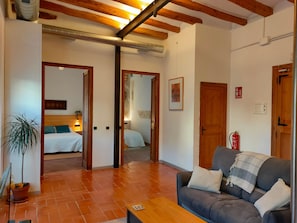 Una sala de estar con un sofá, una mesa de centro y una planta en maceta. Se observan vigas de madera en el techo y un extintor en la pared.