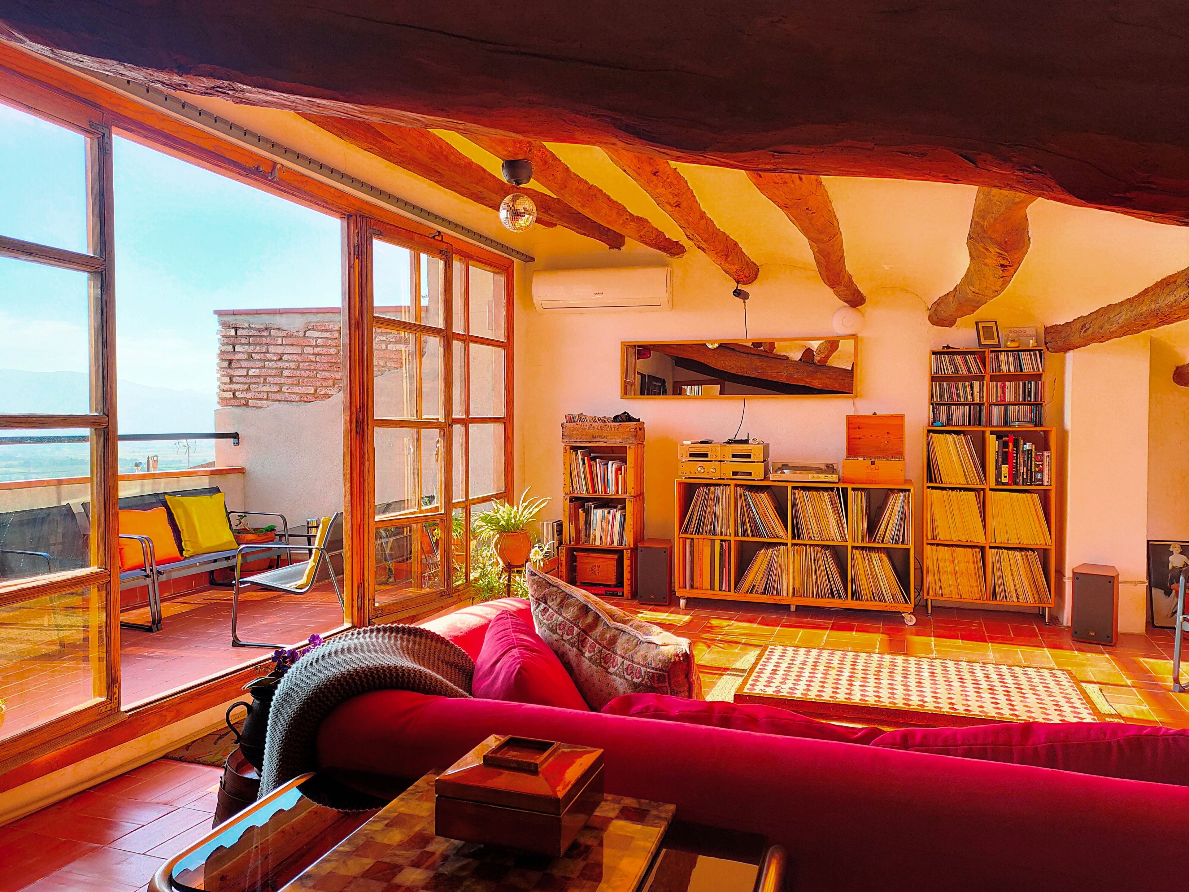 Un soggiorno accogliente con una grande finestra, una libreria piena di libri e dischi, un divano rosso e un soffitto in legno.