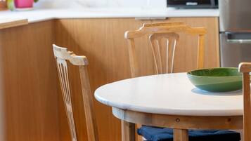 Una silla de comedor de madera con un cojín blanco, un bol verde sobre una mesa blanca, y una cocina al fondo.