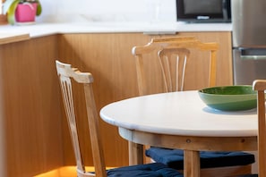 Una silla de comedor de madera con un cojín blanco, un bol verde sobre una mesa blanca, y una cocina al fondo.