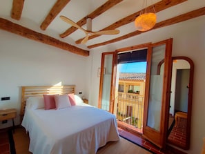 Un dormitorio con techo de madera, una cama con ropa blanca, una mesita de noche de madera y un balcón con vistas a un edificio.