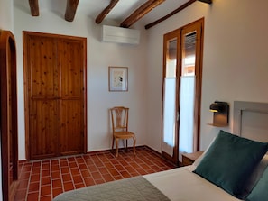 A room with a bed, a wooden wardrobe, a chair, a wall-mounted air conditioner, and a window with blinds.