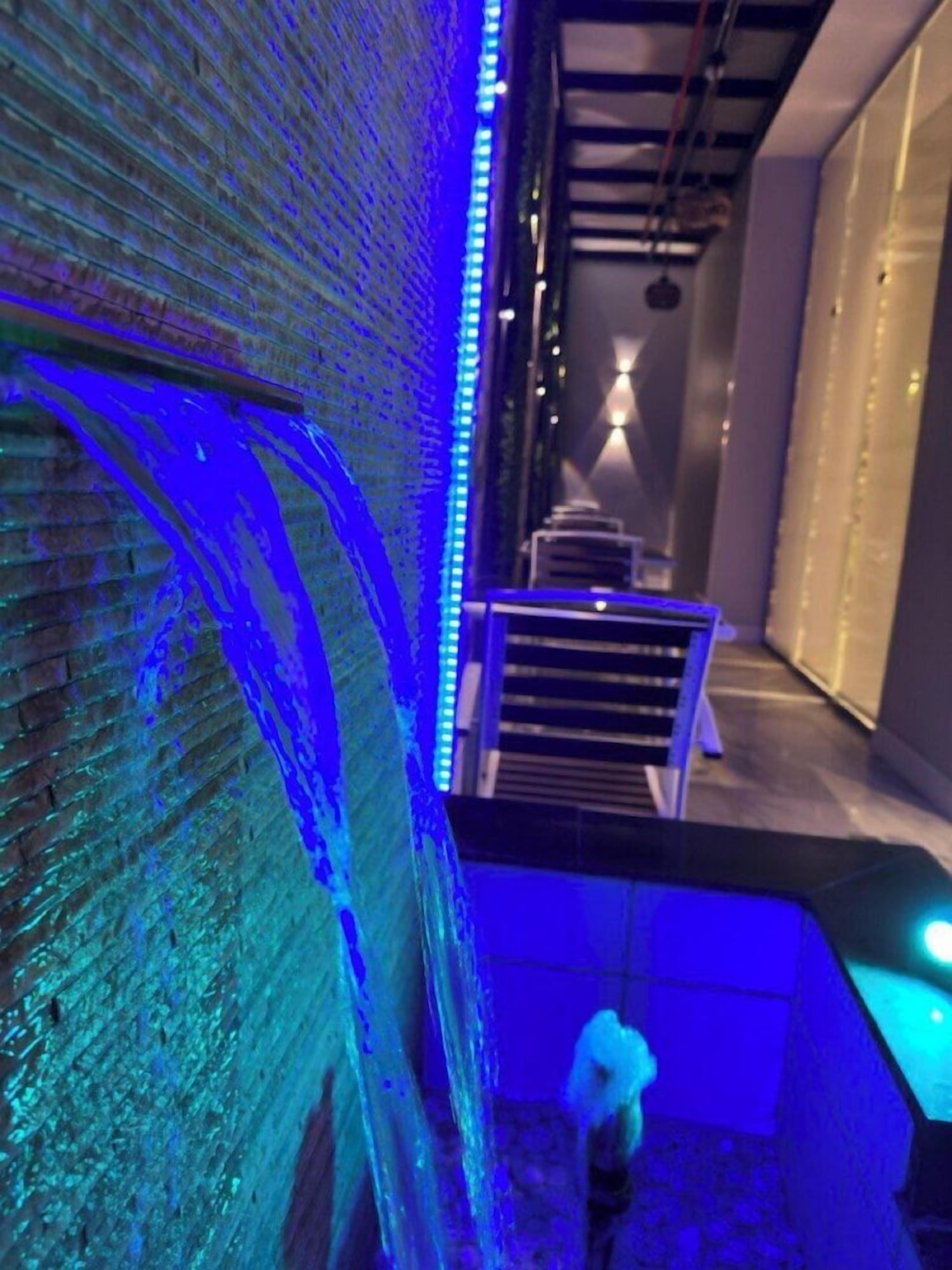 A narrow corridor with a brick wall on one side and a staircase on the other, illuminated by blue lighting.
