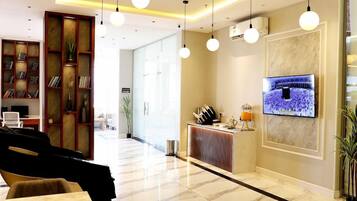 A hotel lobby with a reception desk, a television screen, a seating area with a guitar, and a bookshelf.