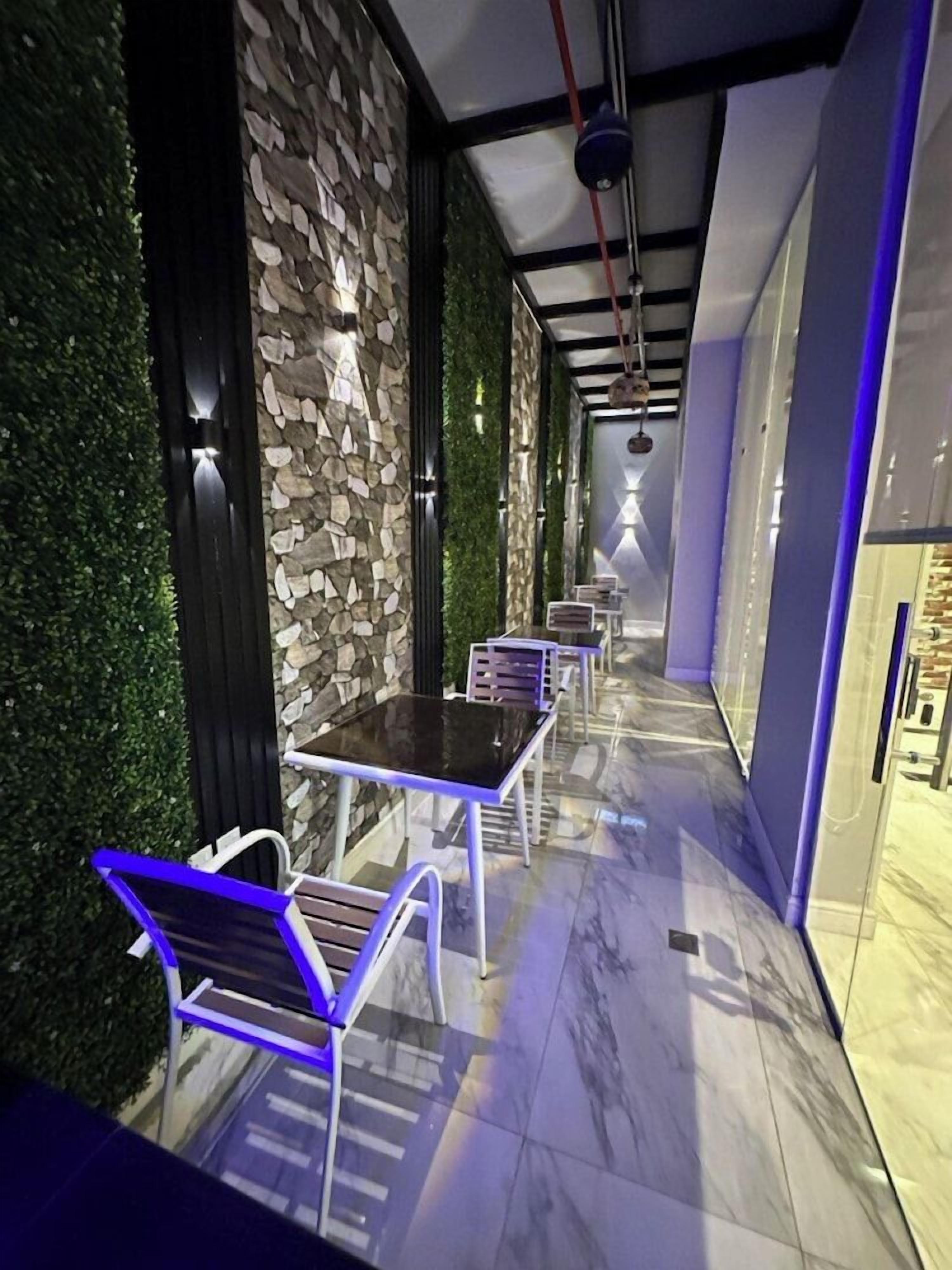 A corridor with a stone wall, green foliage, and a series of tables and chairs.