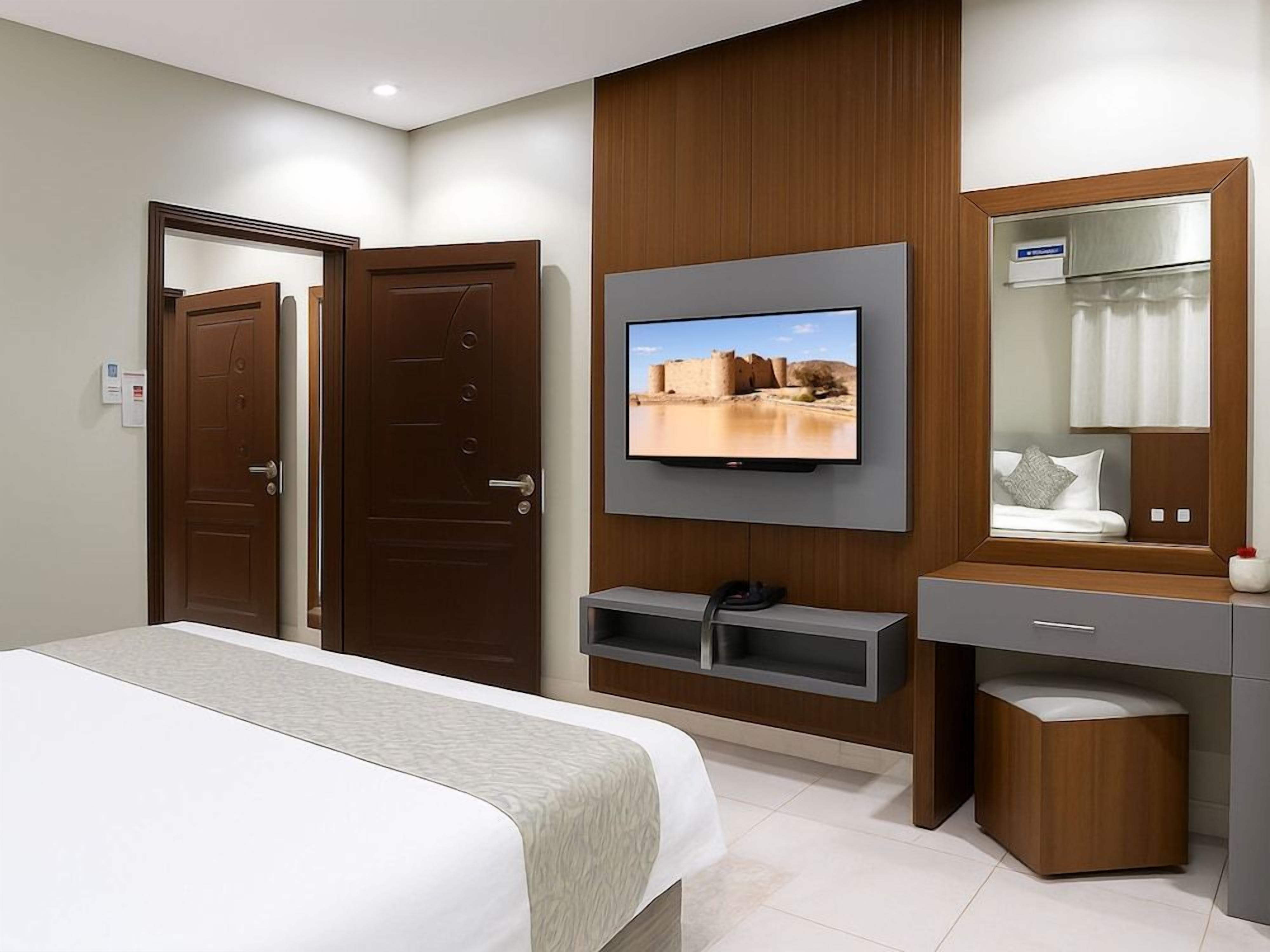 A hotel room with a bed, a TV mounted on the wall, a vanity with a mirror, and a wooden cabinet.