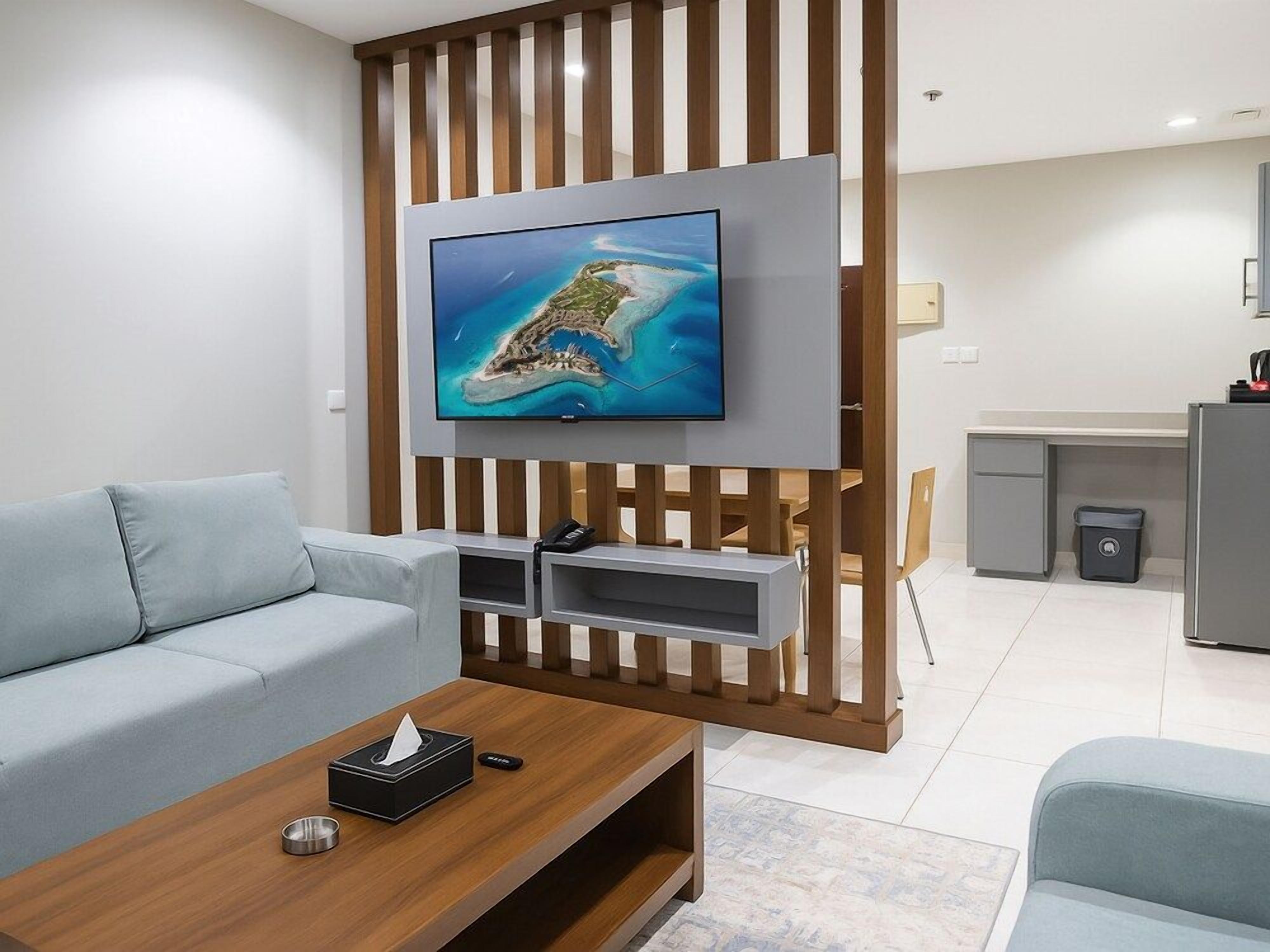 A modern living room with a flat-screen TV mounted on a wooden panel, a light blue sofa, a wooden coffee table, and a kitchen area in the background.