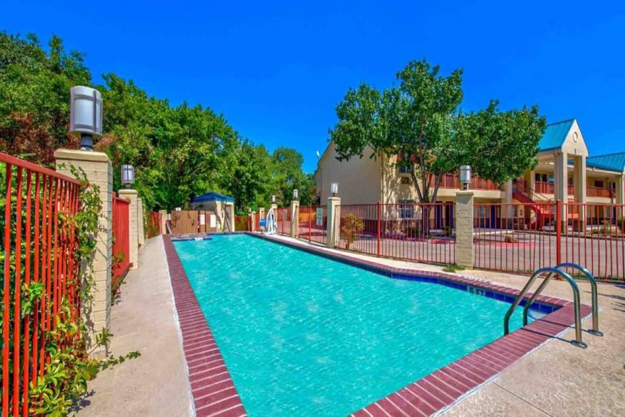 A swimming pool with a red fence, a covered area, and a building with a blue roof. - 2 QUEEN BEDS | MOB ACC TUB NSMK MICFRIDG (ARLINGTON)