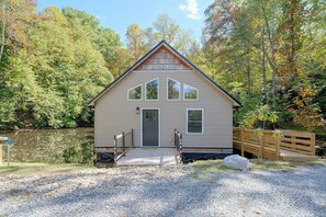 Une cabane avec une véranda à l’avant, une petite terrasse et un étang en arrière-plan.