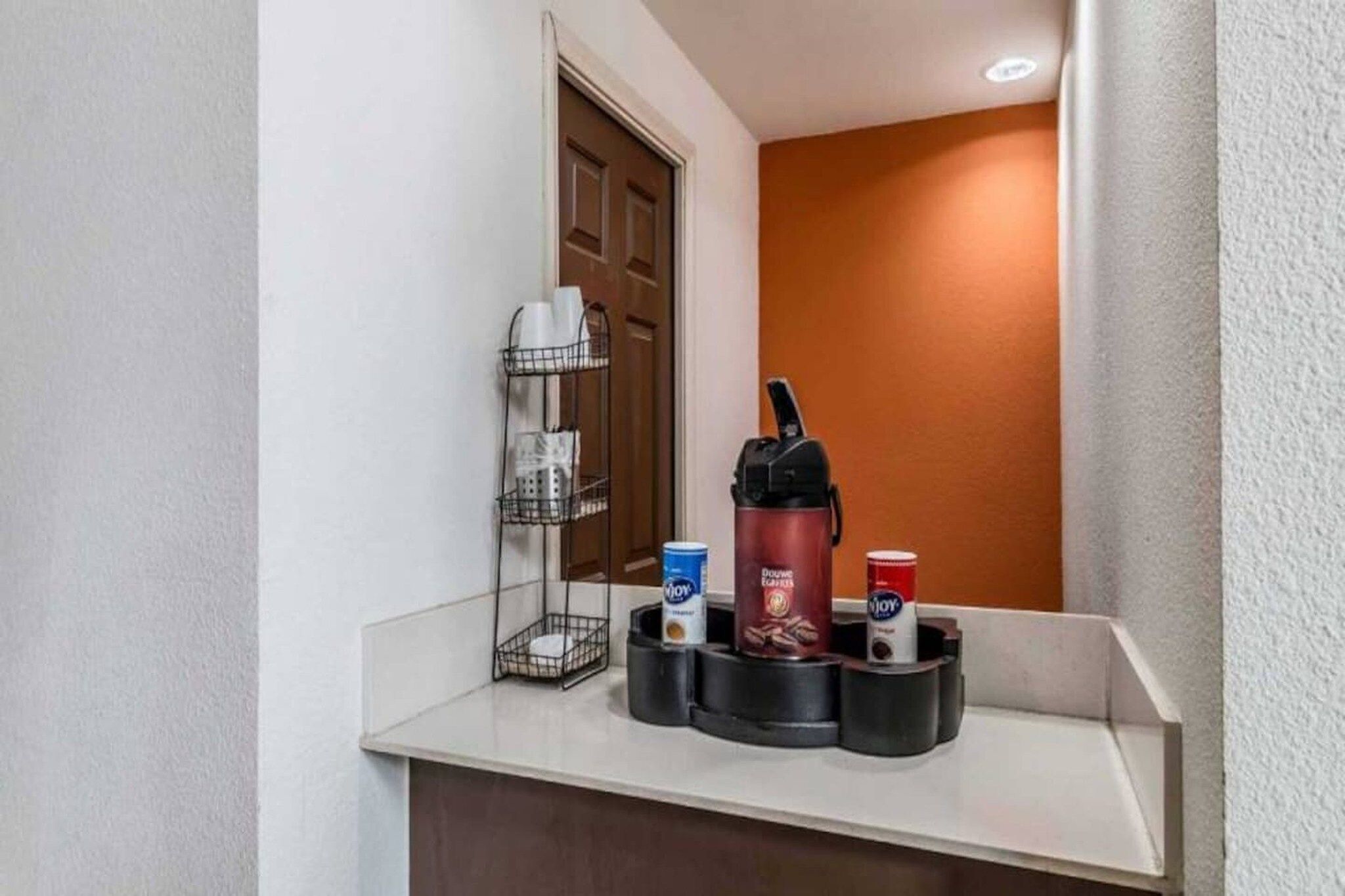 Zona de recepción de hotel con estación de café, cafetera, portavasos y un pequeño cubo de basura.