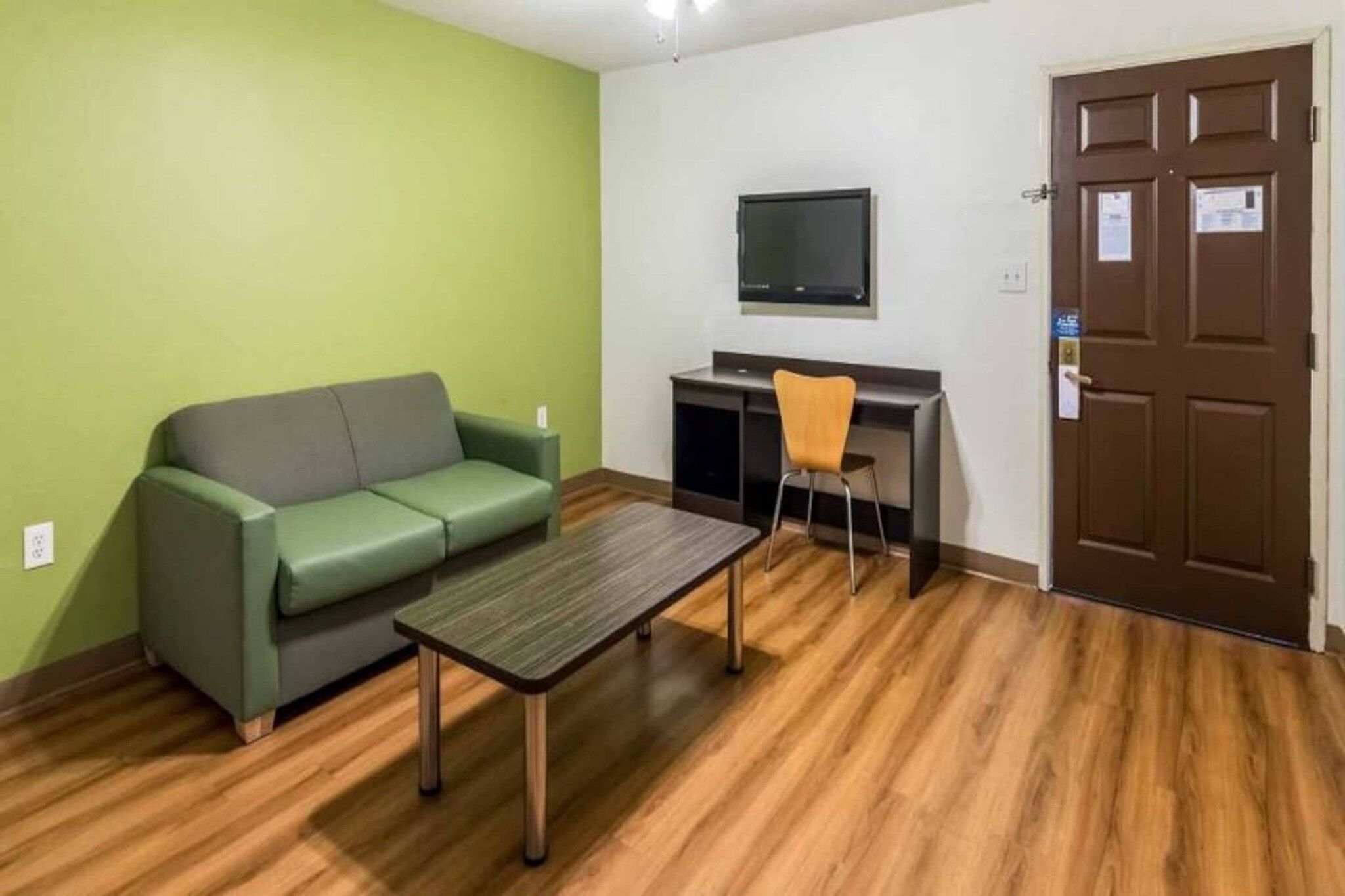 A room with a green sofa, a wooden table, a desk with a chair, a television mounted on the wall, and a brown door.