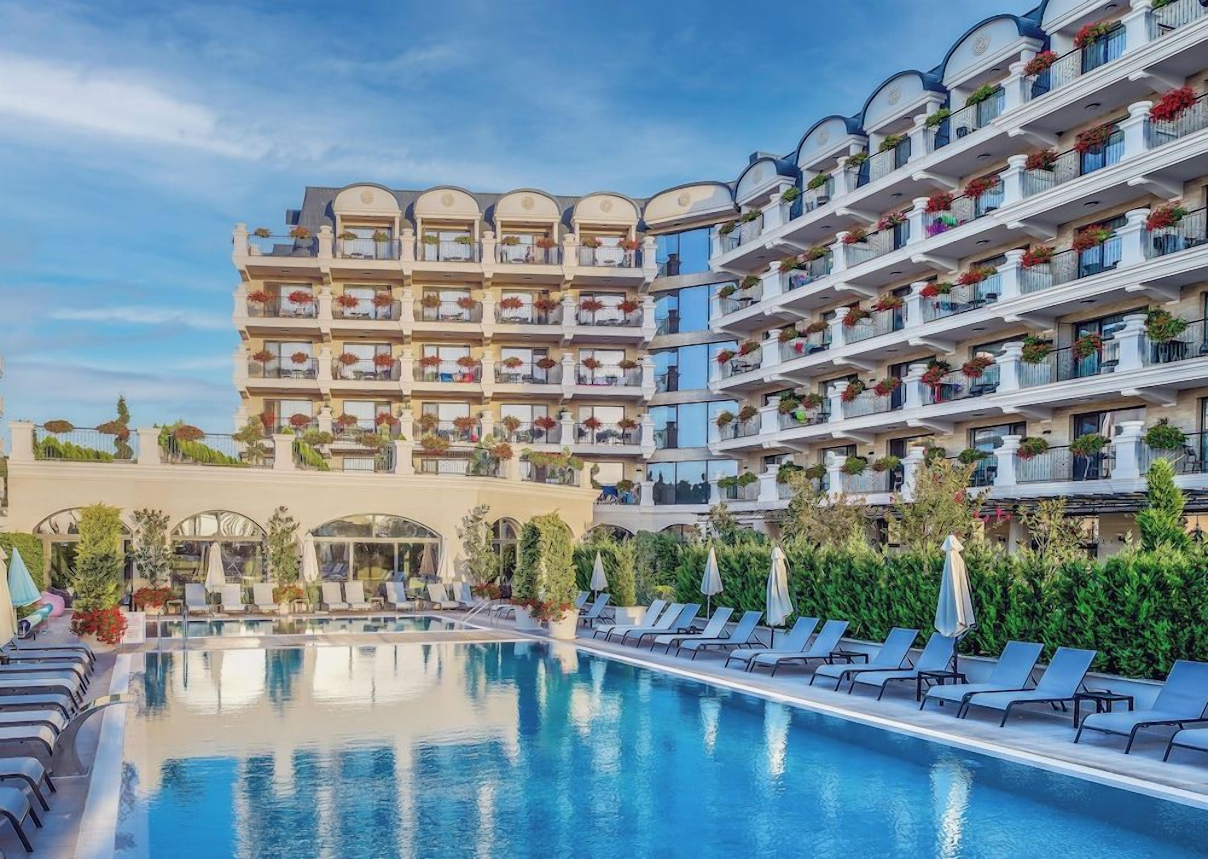 A hotel with a swimming pool, multiple balconies with plants, and a clear sky.