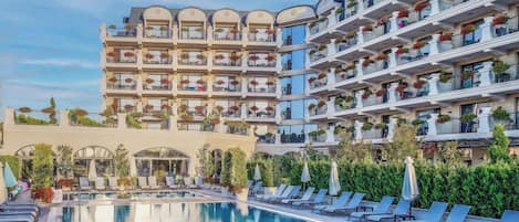 A hotel with a swimming pool, multiple balconies with plants, and a clear sky.