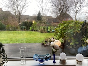Een raam met uitzicht op een tuin, een glazen vaas met bloemen, een blauwe glazen schaal en een glazen karaf.