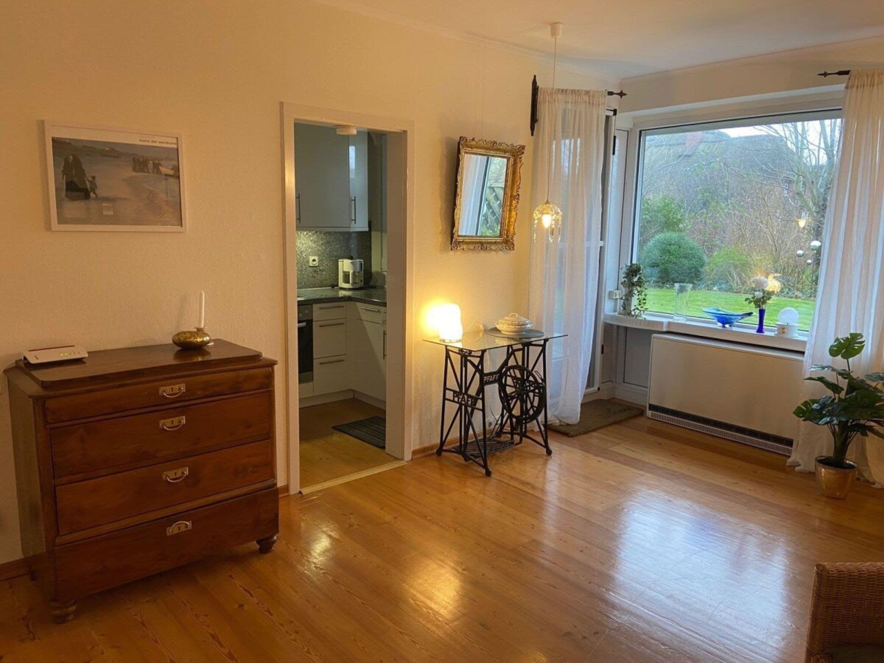 Une pièce avec une commode en bois, une table avec une lampe, une fenêtre avec des rideaux et une vue sur un jardin.