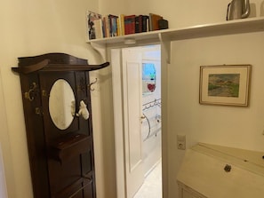Een gang met een houten deur, een witte muur, een plank met boeken, een ingelijst beeld en een witte kast.