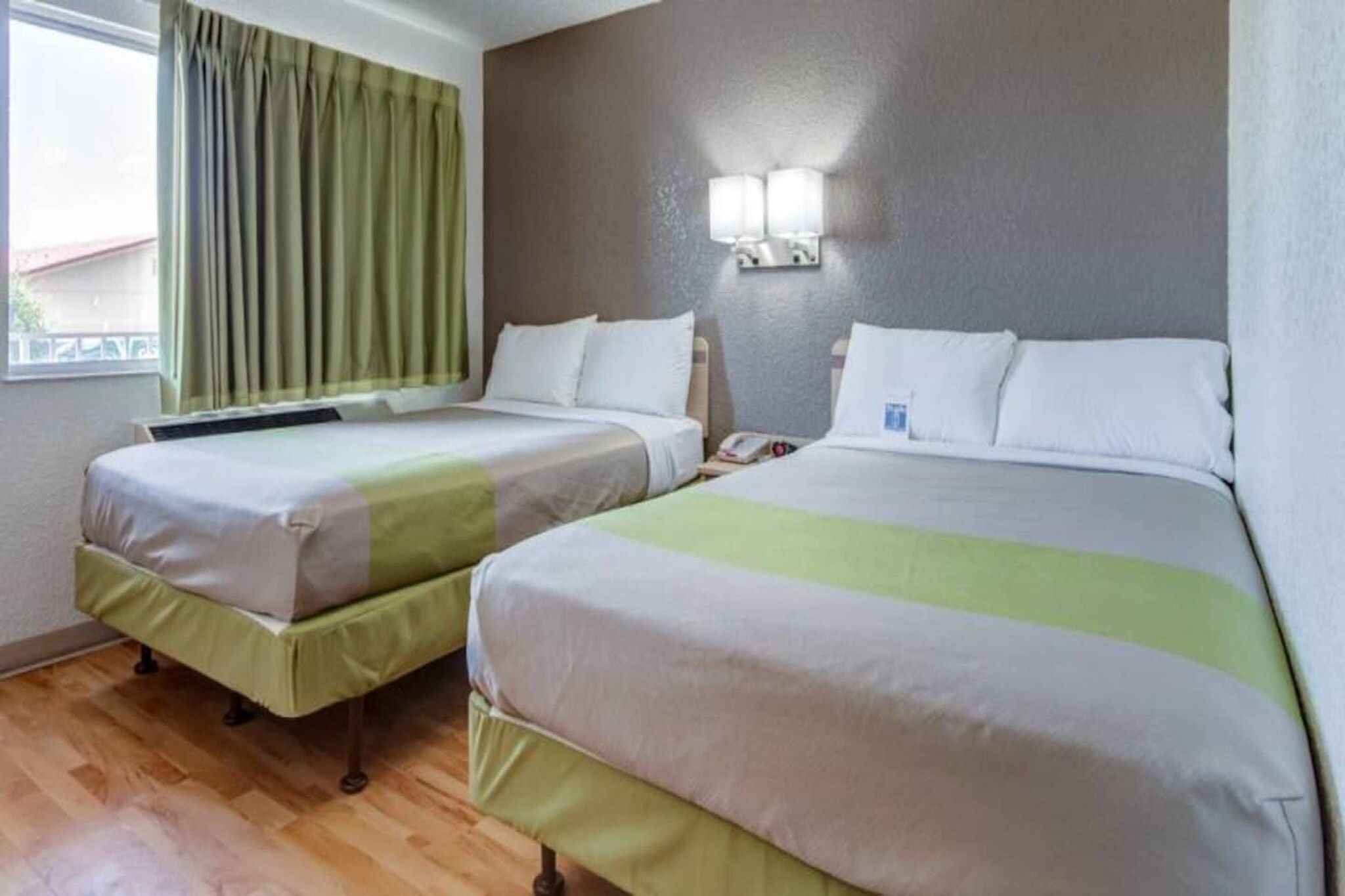 Una habitación de hotel con dos camas, una ventana con cortinas, una lámpara de pared y suelo de madera.