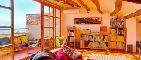 Una sala de estar acogedora con un ventanal grande, una estantería llena de libros y discos, un sofá rojo y un techo de madera.