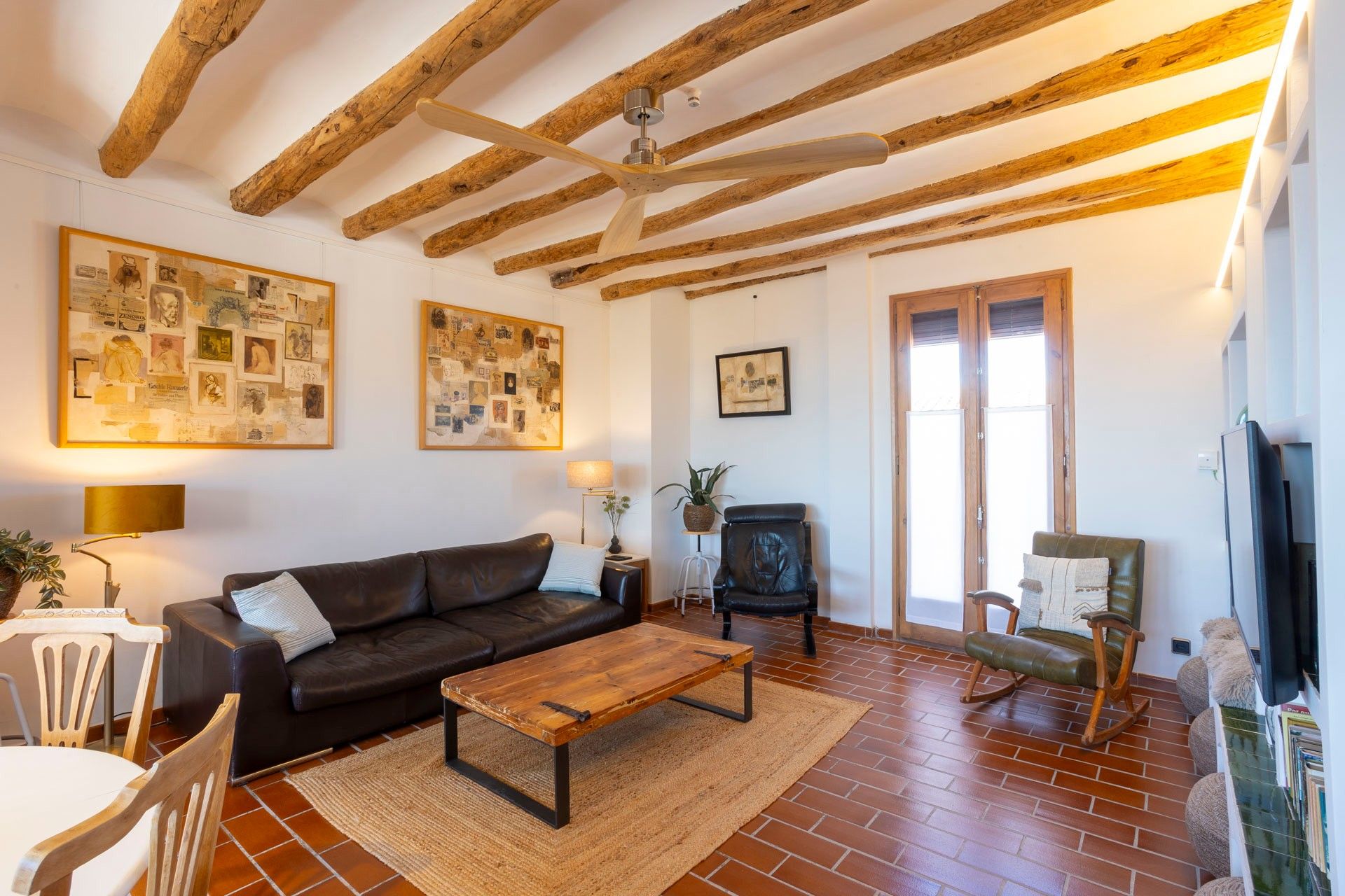 Sala de estar com vigas de madeira aparente, um sofá de couro marrom, mesa de centro de madeira e uma televisão em um suporte.