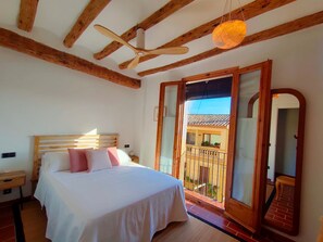 Un dormitorio con techo de madera, una cama con ropa blanca, una mesita de noche de madera y un balcón con vistas a un edificio.
