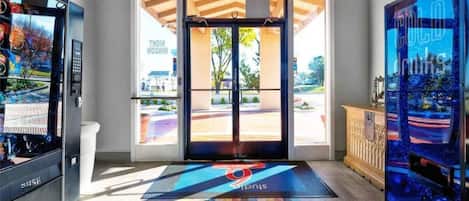 A hotel entrance with a modern design, including a reception desk, a digital screen, and a colorful mat with the hotel logo.