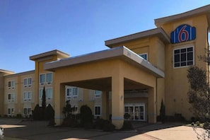 A multi-story hotel with a prominent '6' sign, a covered entrance, and a parking lot. - 1 KING BED | MOBIL ACCESS TUB NSMK MICFRIDG (JOSHUA)