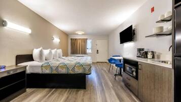A compact hotel room with a bed, a kitchenette, a TV, and a window with curtains.