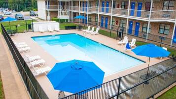 A swimming pool with lounge chairs and umbrellas, surrounded by a fenced area with a building in the background.
