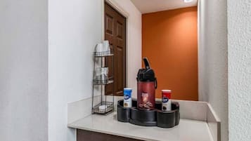 Zona de recepción de hotel con estación de café, cafetera, portavasos y un pequeño cubo de basura.