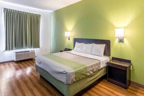 A hotel room with a bed, bedside tables, a nightstand, a lamp, a window with curtains, and an air conditioning unit. - Comfy 1 King Bed | Non-Smoking (NORTH RICHLAND HILLS)