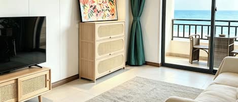 Una sala moderna con televisor de pantalla plana, un mueble con varios cajones, un cuadro abstracto y colorido, y vista al mar a través de una puerta corrediza de vidrio.
