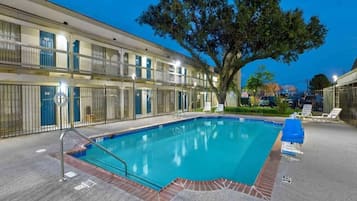 A swimming pool with a tiled deck, surrounded by a fence, and a large tree in the background.