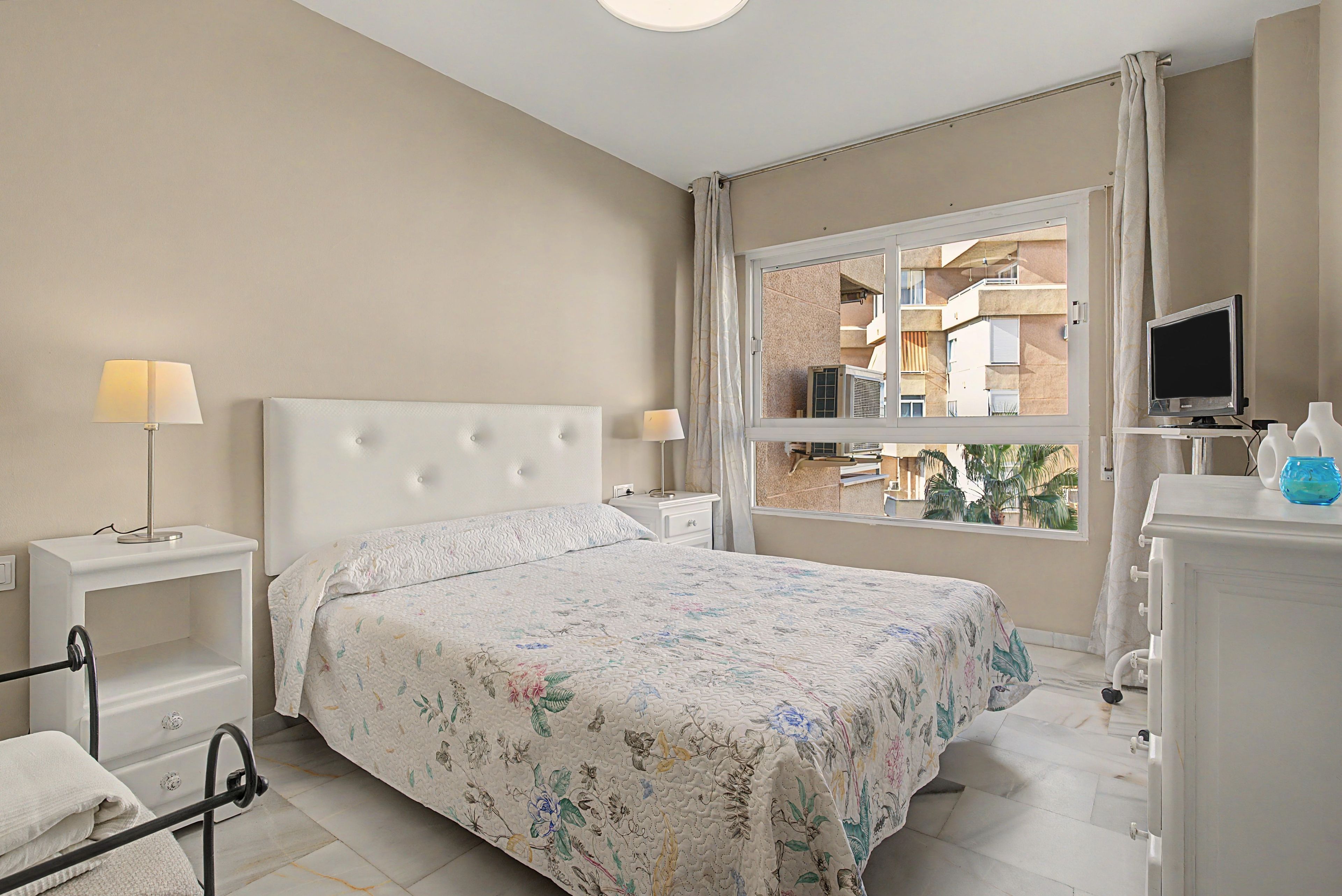 A hotel room with a bed, two bedside tables, a TV, and a window with a view of buildings.