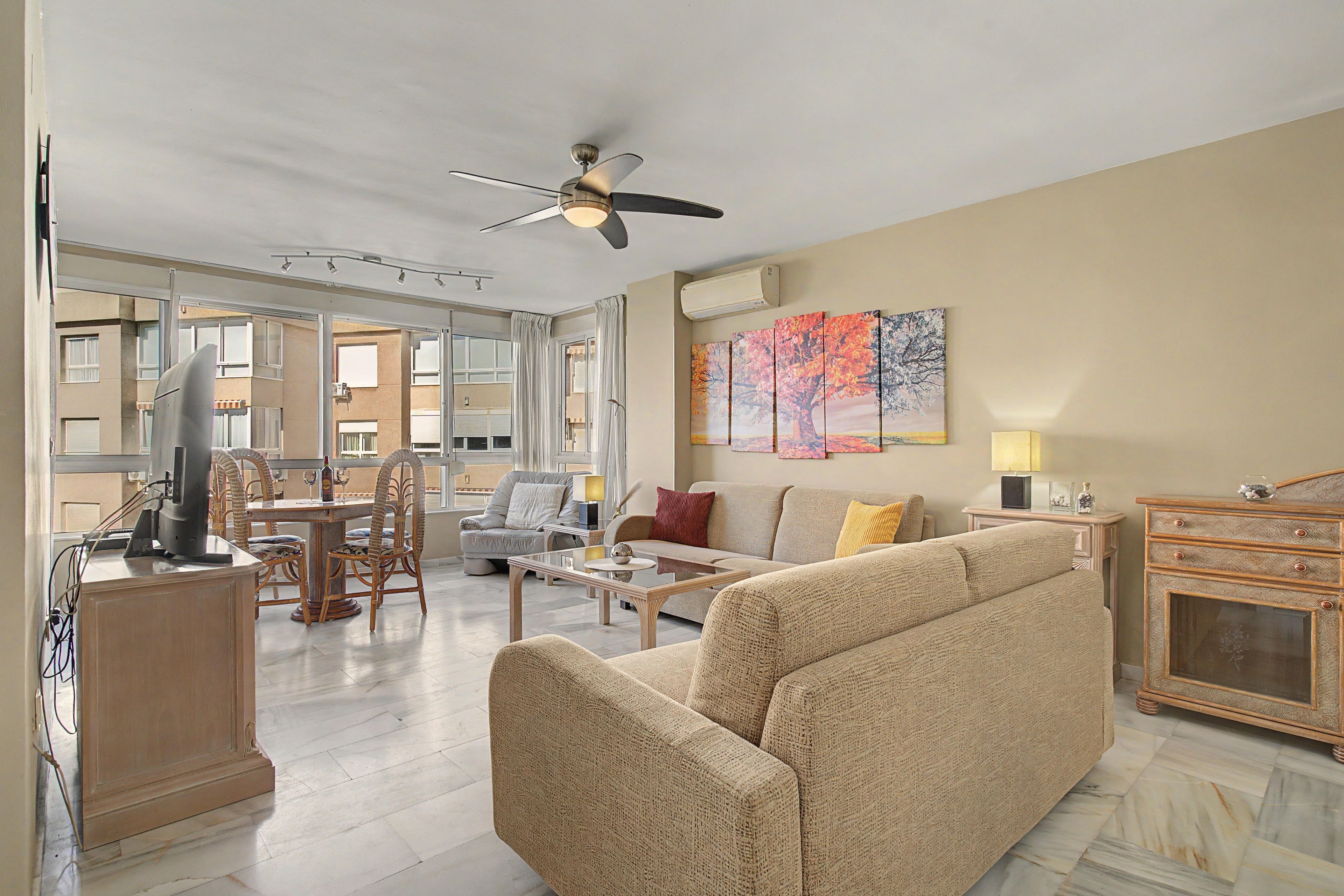A spacious living room with a high ceiling, a ceiling fan, a large window, a sofa, a coffee table, and a television.