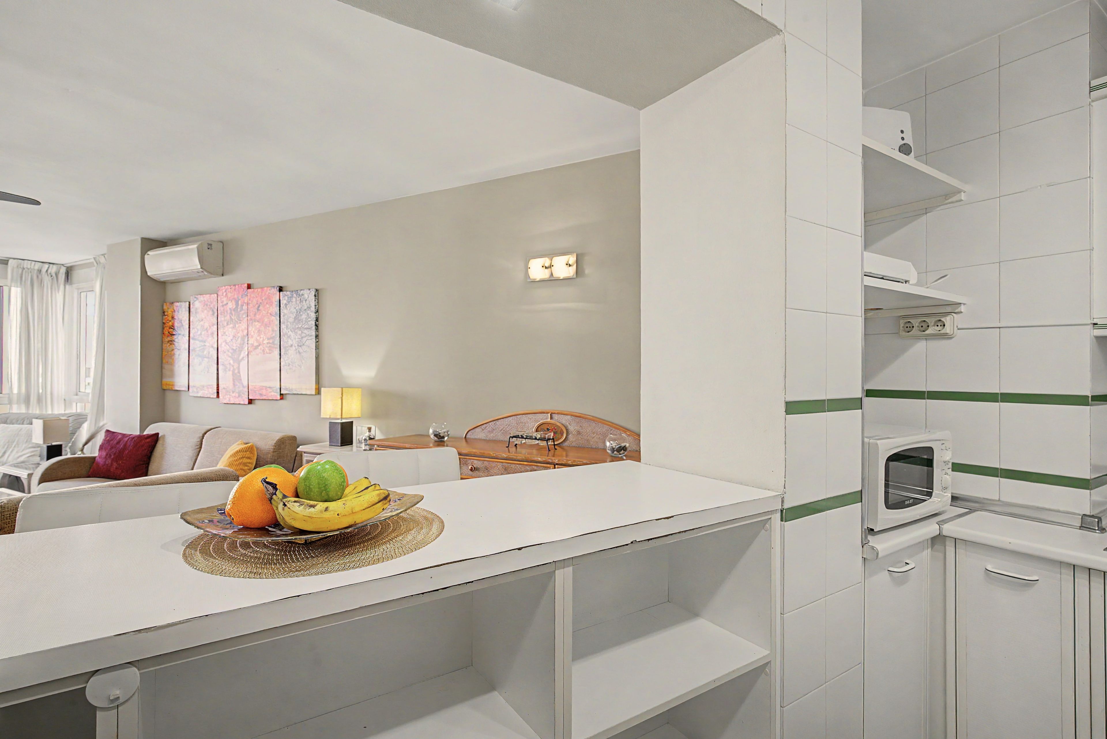 A modern hotel room with a kitchenette, a dining area with a fruit bowl, a bed, and a view of the city.