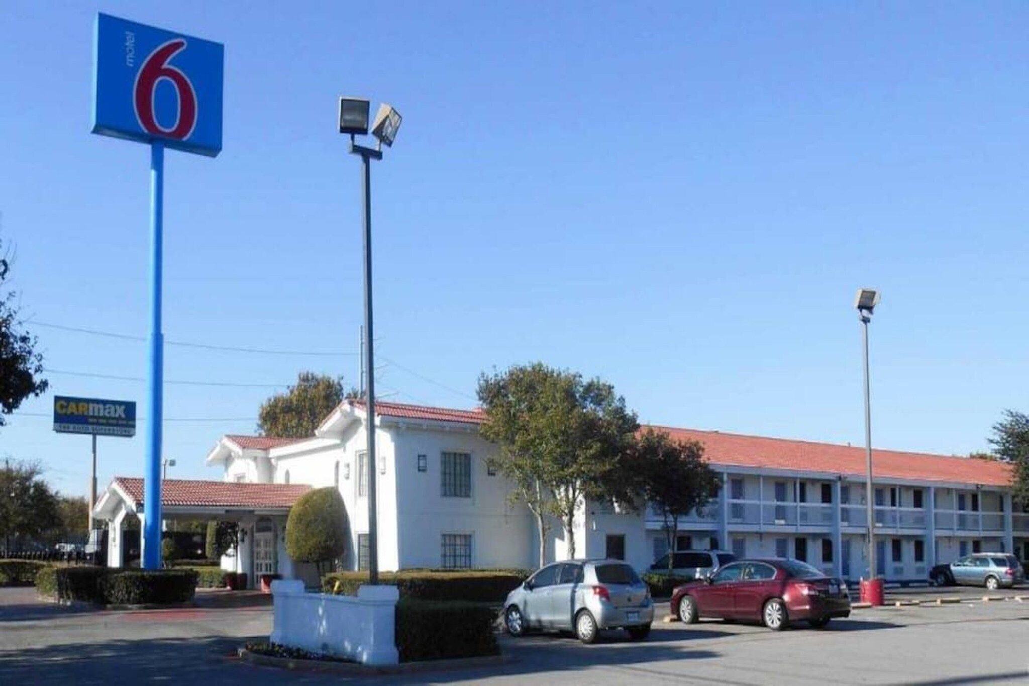 A motel with a red roof, a blue sign with the number 6, and a parking lot with several cars.