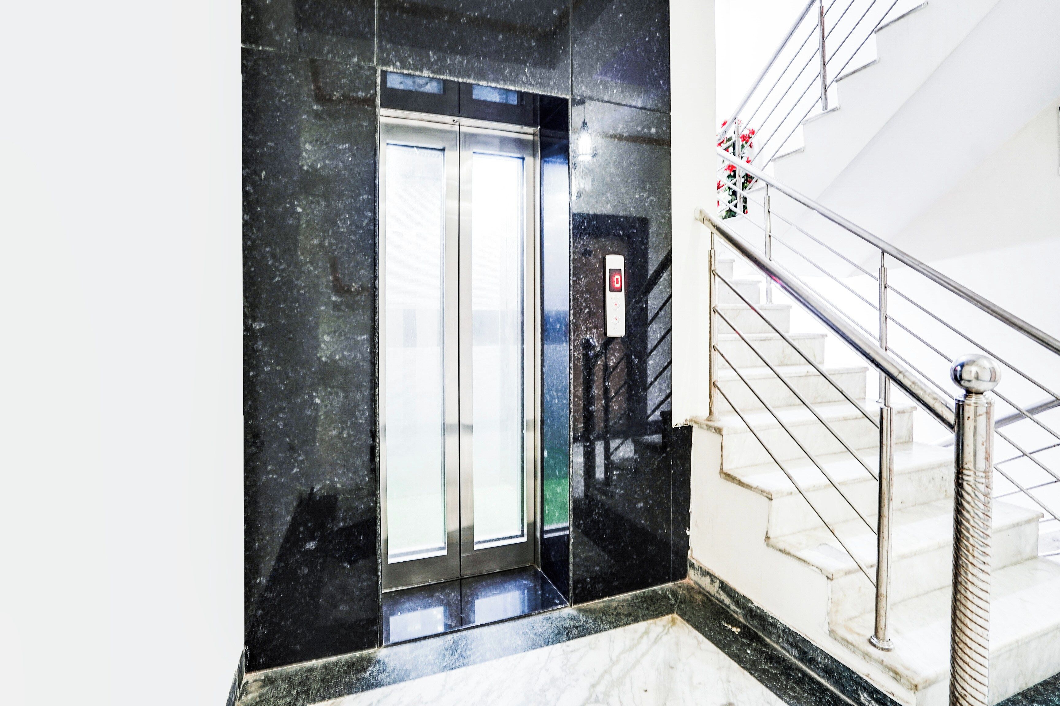 Un ascensor moderno con pantalla digital, piso de mármol y escalera con barandillas de metal.