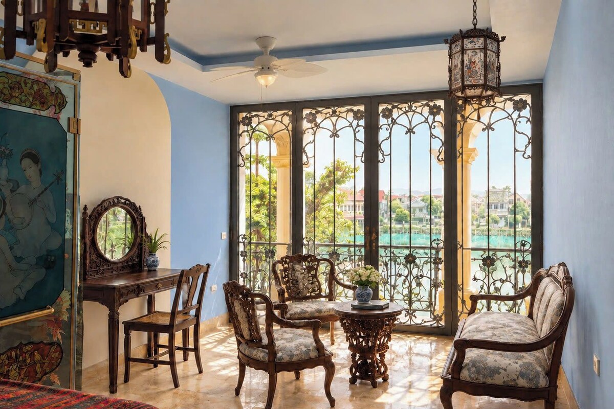 A room with a balcony, a ceiling fan, a table with chairs, and a view of a body of water and greenery.