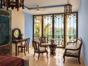 A room with a balcony, a ceiling fan, a table with chairs, and a view of a body of water and greenery. - Thiên Ý Lake Hotel (Phu Xuan)