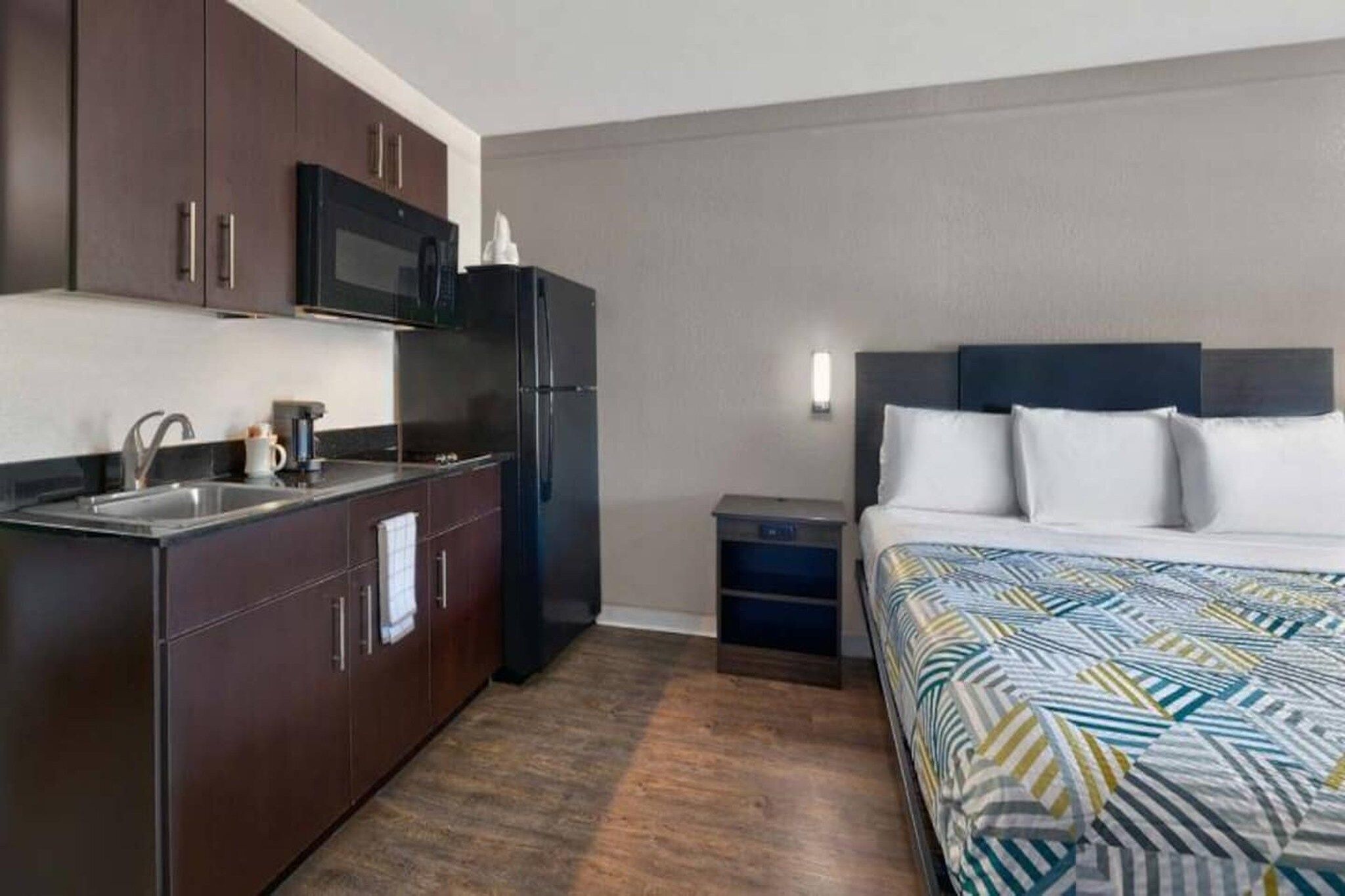 A compact hotel room with a kitchenette, a bed with patterned bedding, and a small bedside table.