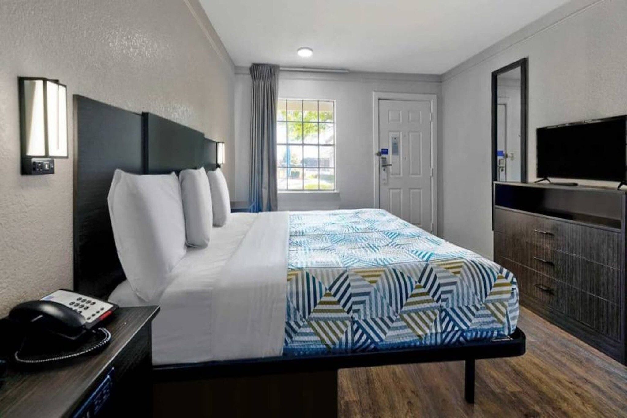A hotel room with a bed, bedside table with a telephone, a desk with a television, a wardrobe, and a door with a window view.