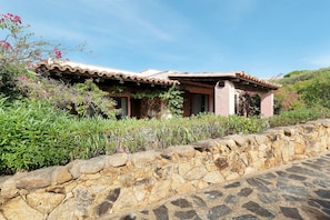 A stone-walled building with a tiled roof, surrounded by lush greenery and flowering plants. - Villa 'Nica' with Private Terrace and Private Garden (Golfo Aranci)